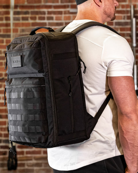 Man wearing large black CONQUER Backpack standing again brick wall