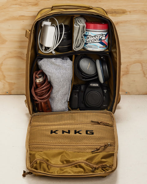 Small desert KNKG CONQUER Backpack open, resting on wooden box, showing internal dividers packed with gym and camera gear