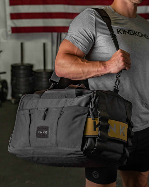 Man wearing black CORE Duffel over shoulder in gym, with American Flag in background