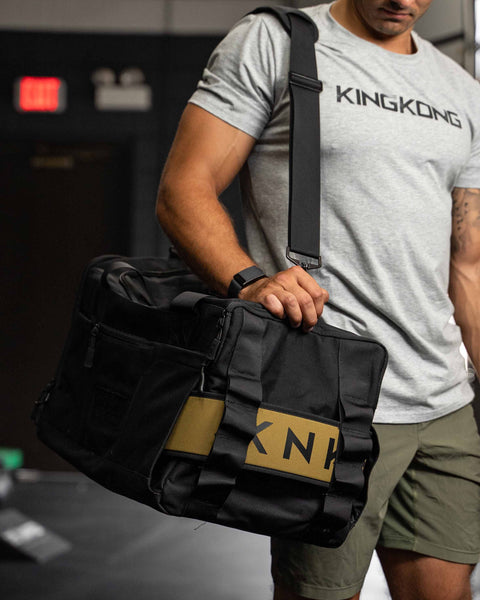 Man wearing large black KNKG CORE Duffel over shoulder, showing lifting belt wrapped around bag, inside sleeve
