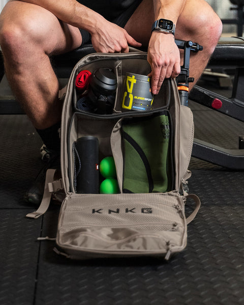A person sitting on a bench with neutral beige KNKG CONQUER backpack open on their lap, displaying various compartments with items like a water bottle and a laptop.