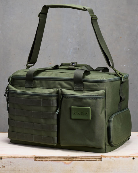 Green KNKG CONQUER Duffel sitting on wooden box with shoulder strap suspended
