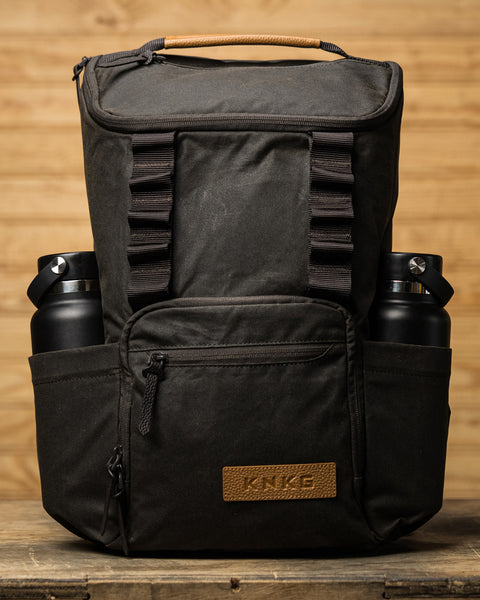 Front view of KNKG Waxed Canvas Backpack sitting on box with drink bottles in side pockets.