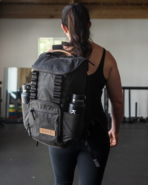 Person wearing KNKG Waxed Canvas Backpack with water bottles in a gym setting