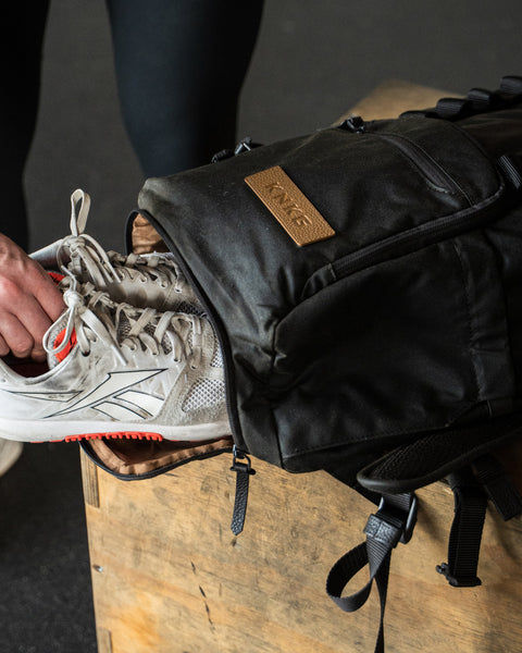 Person putting a white sneaker into KNKG Waxed Canvas Backpack on a wooden surface.