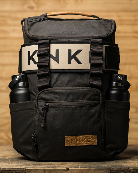 KNKG Waxed Canvas Backpack sitting on wooden box with weightlifting belt and bottles in side pockets