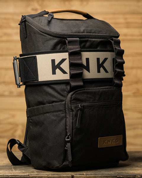 KNKG Waxed Canvas Backpack sitting on wooden box with matching weightlifting belt wrapped around backpack