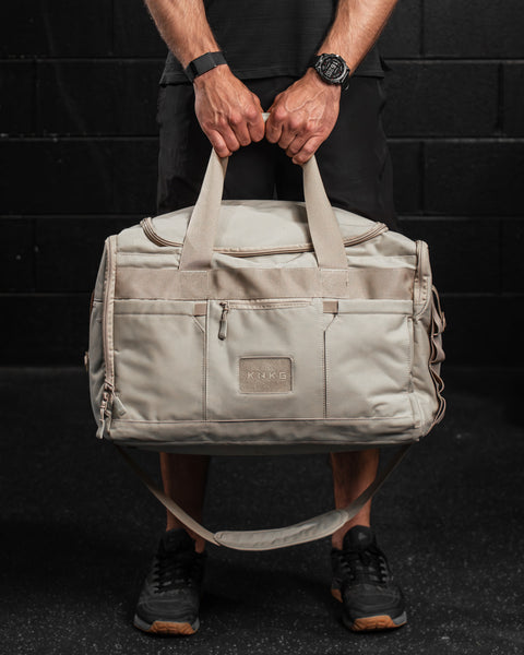 Man holding large neutral beige CORE Duffel by handles, standing against black brick wall