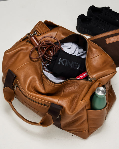 Birds eye view of large tan HERITAGE leather duffel packed with gym gear and NOBULL sneakers in background