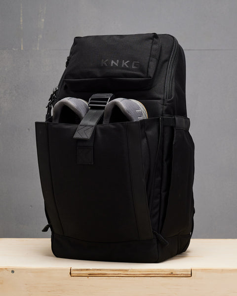 Black KNKG PLUS Backpack with shoes inside of front compartment sitting on wooden box