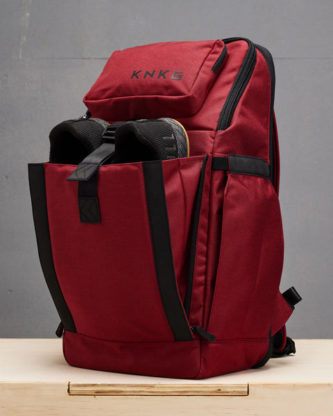 Red KNKG PLUS Backpack sitting on wooden box with shoes inside front compartment