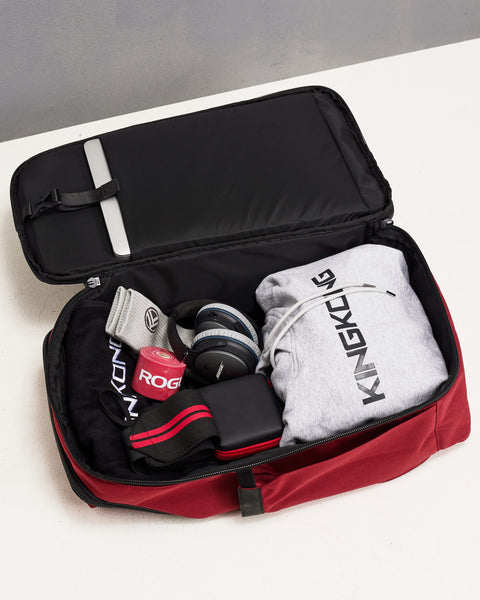 Red KNKG PLUS Backpack opened in clamshell style, laying on floor, packed with hoodie, headphones, wrist wraps and mobility accessories