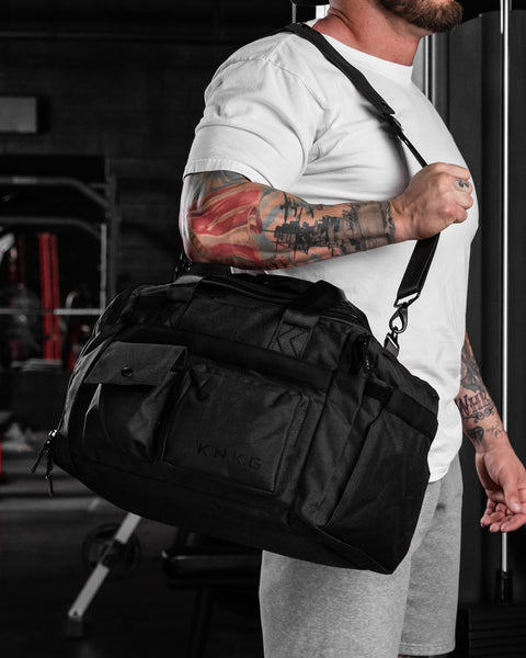 A person carrying small black KNKG PLUS Duffel bag over their shoulder in a gym setting.