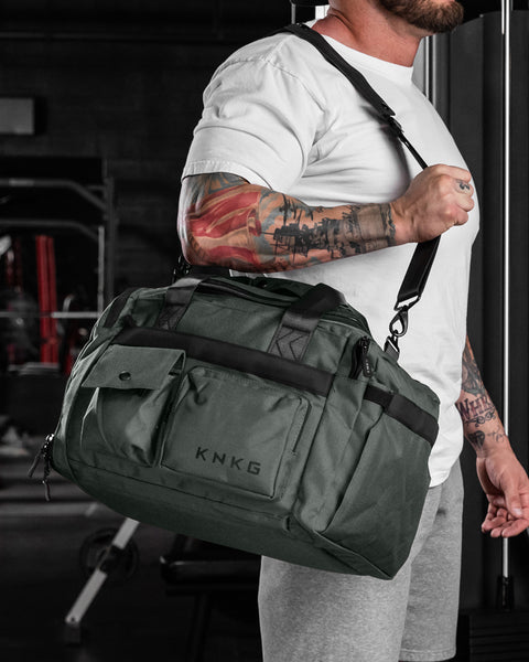 Man wearing charcoal grey KNKG PLUS duffel bag over shoulder inside gym setting