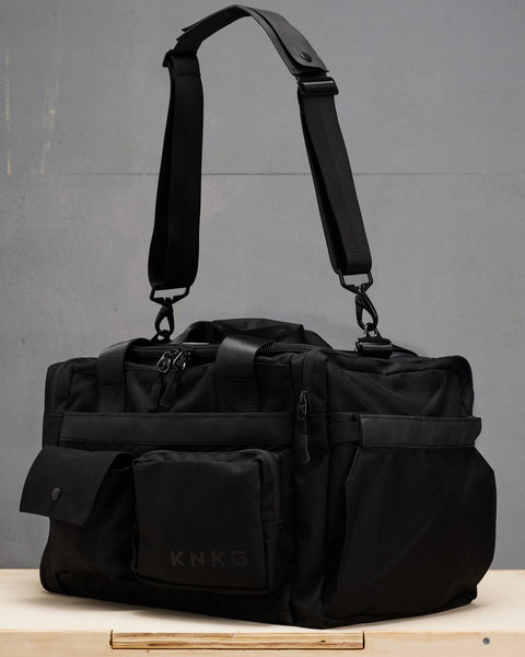 small black KNKG PLUS Duffel bag sitting on wooden box with shoulder strap suspended