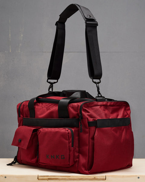 small red KNKG PLUS Duffel sitting on wooden box with shoulder strap suspended