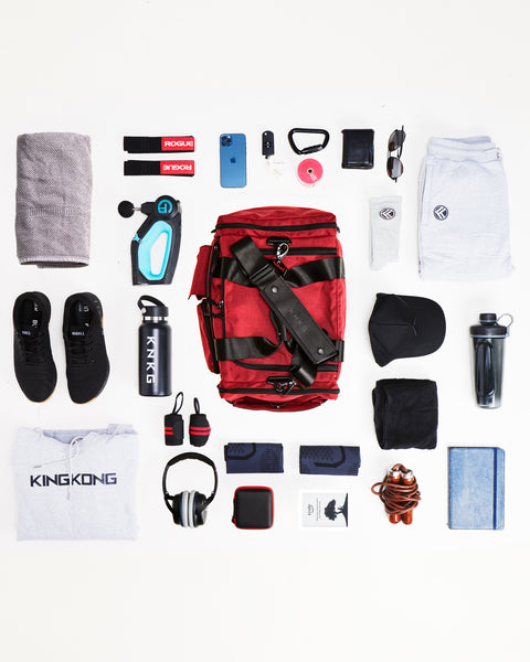 Flat lay of small red KNKG PLUS Duffel sitting on floor, surrounded by gym gear that fits inside bag