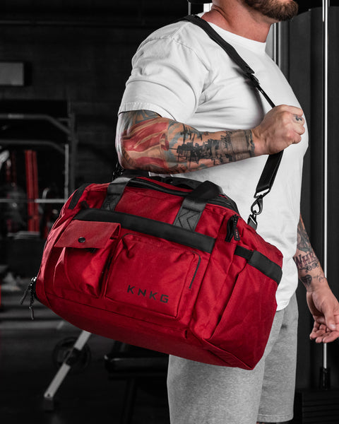 Man wearing small red KNKG PLUS Duffel over shoulder inside dark gym setting