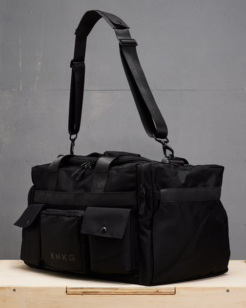 Side view of large black KNKG PLUS duffel bag sitting on wooden box with shoulder strap suspended