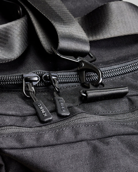 Close up of KNKG branded zippers and metal buckles on black KNKG PLUS duffel bag