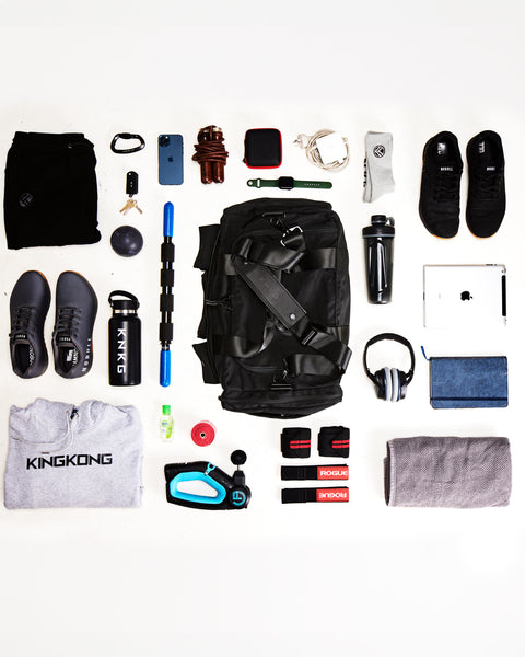Flay lay of large black KNKG PLUS duffel bag sitting on floor, surrounded by sneakers, gym clothes, towel, laptop and fitness and mobility accessories