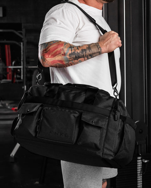A person carrying a large black KNKG PLUS duffel bag over their shoulder in a gym setting.