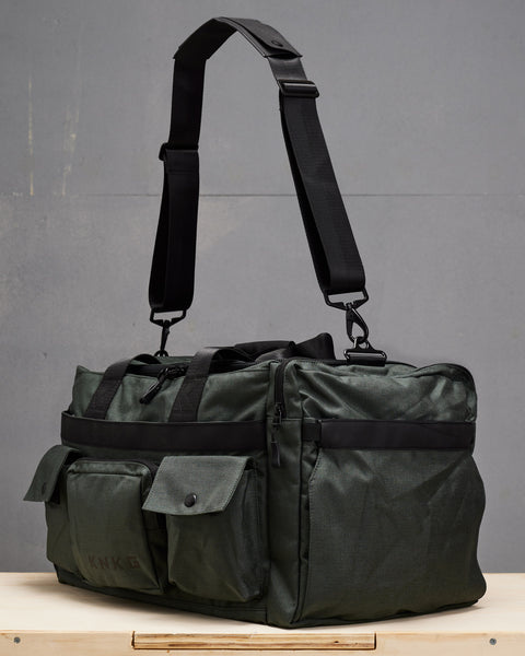 Side view of charcoal grey KNKG PLUS Duffel bag sitting on wooden box, with shoulder strap suspended