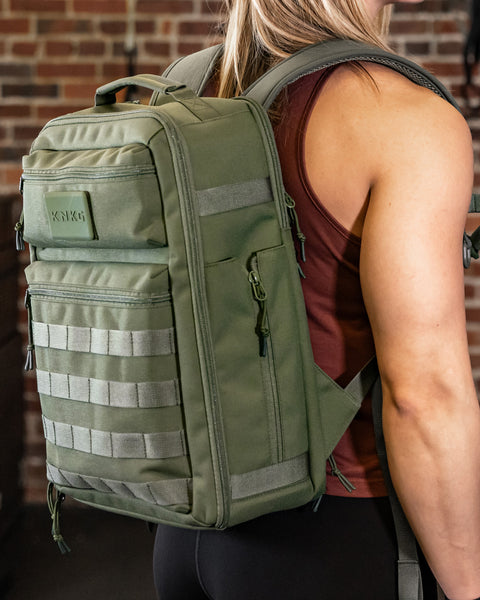 Woman standing against brick wall in gym wearing small green KNKG CONQUER Backpack