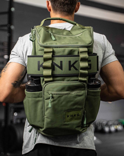 Man wearing green CORE Backpack with matching weight lifting belt wrapped around bag, inside dark gym setting.