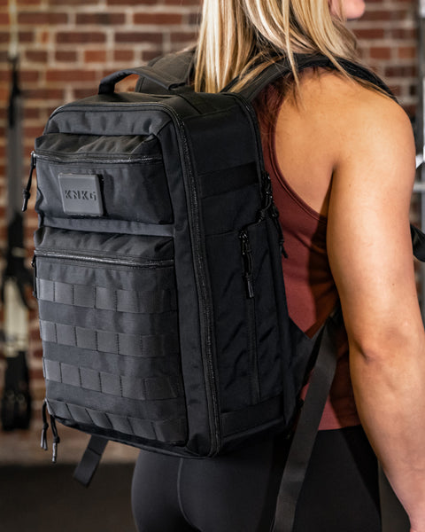 Woman wearing Small black KNKG CONQUER Backpack standing against brick wall inside gym