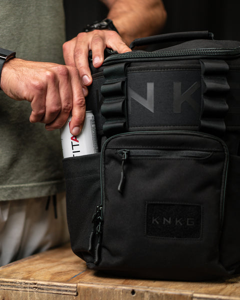 Person reaching for FITAID inside drink bottle pocket of KNKG Mini CORE Backpack in Black