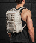 Person wearing a neutral beige mini size KNKG CORE Backpack against a black brick wall.