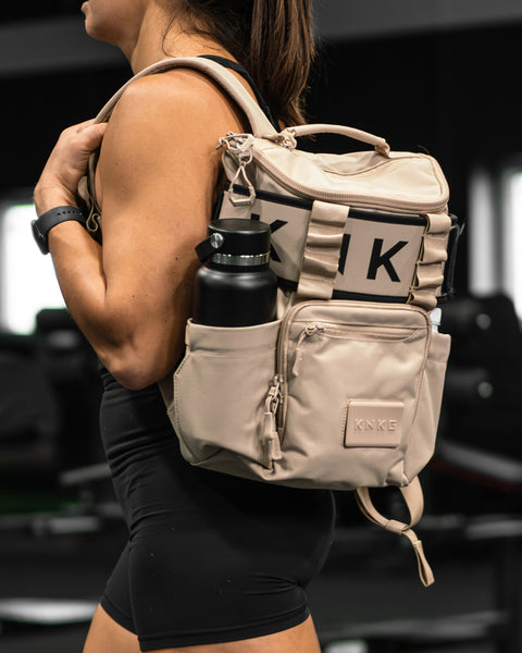 Person wearing neutral beige mini size KNKG CORE Backpack with a water bottle, blurred background