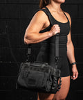 Person wearing KNKG mini black CORE Duffel over shoulder against a black brick wall.