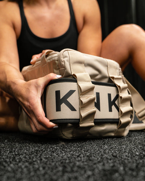 Person sitting on floor of gym reaching for lifting belt attached to Neutral beige KNKG mini CORE Duffel