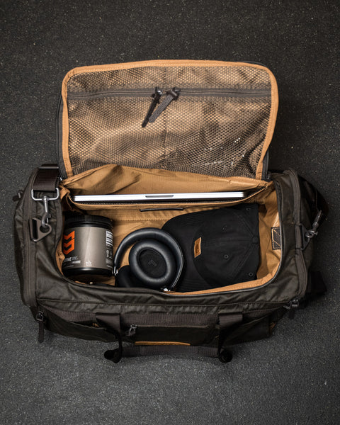 Open KNKG Waxed Canvas Duffel with laptop, coffee mug, and headphones on a dark background