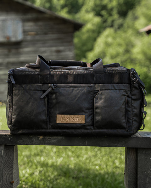 KNKG Waxed Canvas Duffel sitting on wooden bench in front of wooden barn