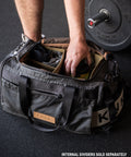 Person grabbing gym essentials out of KNKG Waxed Canvas Duffel sitting on gym floor, with barbell in background