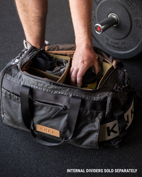 Person grabbing gym essentials out of KNKG Waxed Canvas Duffel sitting on gym floor, with barbell in background