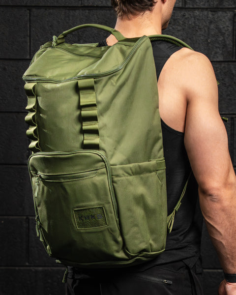 Man standing against black brick wall, wearing large green CORE Backpack